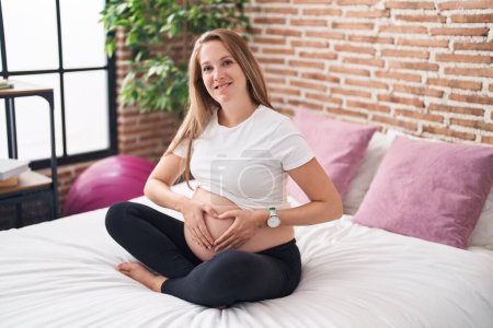 Foto de Mujer embarazada joven sonriendo confiado haciendo gesto de corazón en el vientre en el dormitorio - Imagen libre de derechos