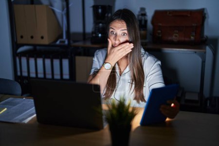Téléchargez les photos : Jeune femme brune travaillant au bureau la nuit choquée couvrant la bouche avec les mains par erreur. concept secret. - en image libre de droit