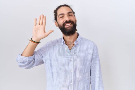 Foto de Hombre hispano con barba usando camisa casual renunciando a decir hola feliz y sonriente, gesto de bienvenida amistoso - Imagen libre de derechos