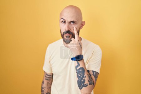 Foto de Hispanic man with tattoos standing over yellow background pointing to the eye watching you gesture, suspicious expression - Imagen libre de derechos