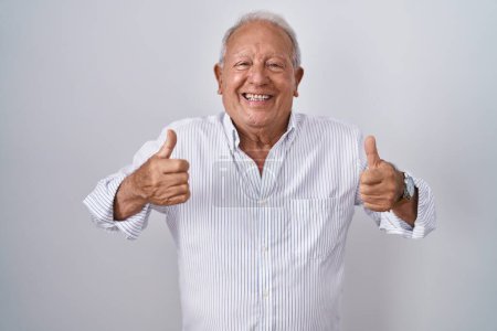 Foto de Senior man with grey hair standing over isolated background success sign doing positive gesture with hand, thumbs up smiling and happy. cheerful expression and winner gesture. - Imagen libre de derechos