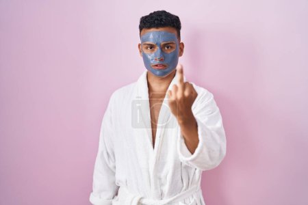Foto de Young hispanic man wearing beauty face mask and bath robe showing middle finger, impolite and rude fuck off expression - Imagen libre de derechos