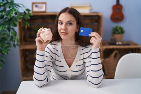 Photo for Young hispanic girl holding piggy bank and credit card relaxed with serious expression on face. simple and natural looking at the camera. - Royalty Free Image