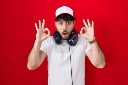 Photo for Hispanic man with beard wearing gamer hat and headphones looking surprised and shocked doing ok approval symbol with fingers. crazy expression - Royalty Free Image