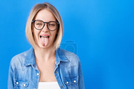 Téléchargez les photos : Young caucasian woman standing over blue background sticking tongue out happy with funny expression. emotion concept. - en image libre de droit