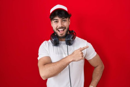 Foto de Hombre hispano con barba con sombrero de gamer y auriculares con una gran sonrisa en la cara, apuntando con el dedo de la mano hacia un lado mirando a la cámara. - Imagen libre de derechos