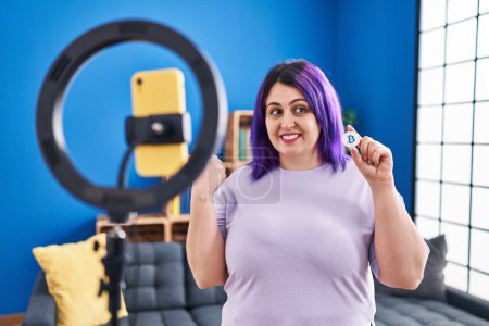 Téléchargez les photos : Plus taille femme avec cheveux violets enregistrement tutoriel Bitcoin avec smartphone à la maison pointant pouce vers le côté souriant heureux avec bouche ouverte - en image libre de droit