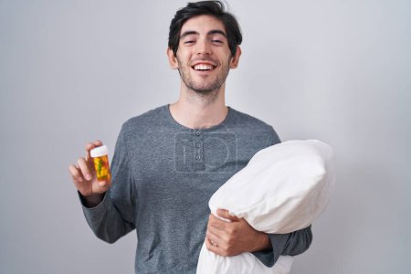 Foto de Joven hombre hispano usando pijama abrazando almohada sosteniendo pastillas sonriendo y riendo en voz alta porque gracioso chiste loco. - Imagen libre de derechos
