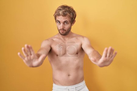 Téléchargez les photos : Homme caucasien debout torse nu portant un écran solaire faisant geste d'arrêt avec les paumes des mains, expression de colère et de frustration - en image libre de droit