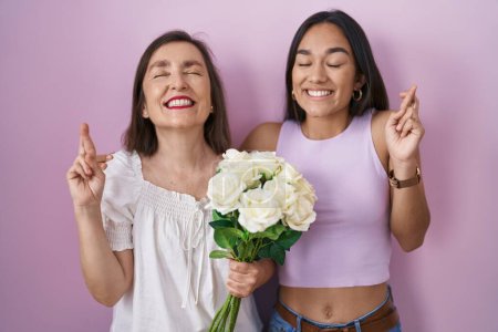 Photo for Hispanic mother and daughter holding bouquet of white flowers gesturing finger crossed smiling with hope and eyes closed. luck and superstitious concept. - Royalty Free Image