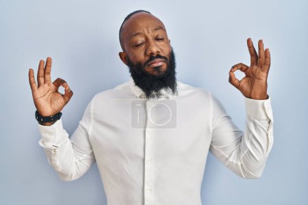 Téléchargez les photos : Homme afro-américain debout sur fond bleu détendu et souriant avec les yeux fermés faisant geste de méditation avec les doigts. concept de yoga. - en image libre de droit