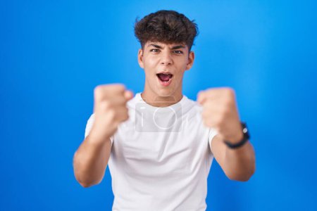 Photo for Hispanic teenager standing over blue background angry and mad raising fists frustrated and furious while shouting with anger. rage and aggressive concept. - Royalty Free Image