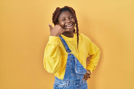 Foto de Mujer africana de pie sobre fondo amarillo sonriendo haciendo gesto telefónico con la mano y los dedos como hablando por teléfono. comunicar conceptos. - Imagen libre de derechos
