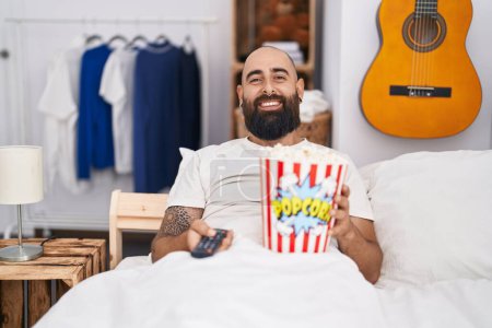 Photo for Young hispanic man with beard and tattoos eating popcorn in the bed smiling with a happy and cool smile on face. showing teeth. - Royalty Free Image