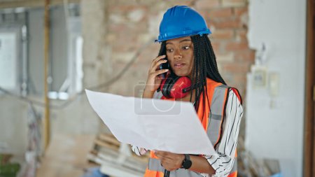Téléchargez les photos : African american woman builder lecture projet de maison parler par smartphone sur le site de construction - en image libre de droit