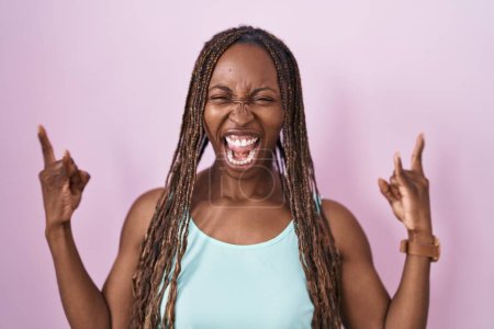 Foto de Mujer afroamericana de pie sobre fondo rosa gritando con expresión loca haciendo símbolo de roca con las manos arriba. Estrella de la música. concepto de música pesada. - Imagen libre de derechos