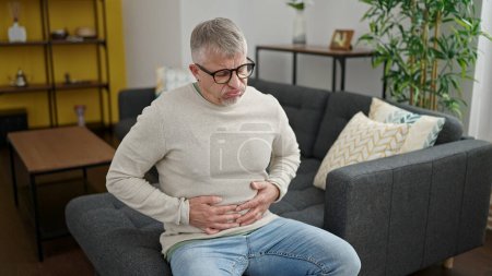 Foto de Hombre de pelo gris de mediana edad que sufre de dolor de estómago sentado en el sofá en casa - Imagen libre de derechos