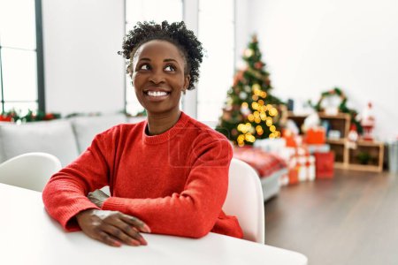 Foto de Mujer afroamericana sonriendo confiada sentada en la mesa junto al árbol de Navidad en casa - Imagen libre de derechos