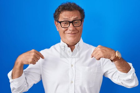 Foto de Hombre hispano de mediana edad de pie sobre fondo azul mirando confiado con sonrisa en la cara, señalándose con los dedos orgullosos y felices. - Imagen libre de derechos