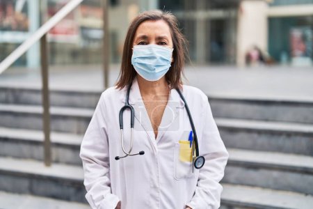 Téléchargez les photos : Femme d'âge moyen portant un uniforme de médecin et un masque médical debout dans la rue - en image libre de droit