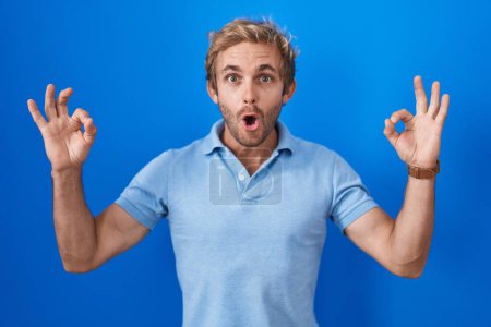 Photo for Caucasian man standing over blue background looking surprised and shocked doing ok approval symbol with fingers. crazy expression - Royalty Free Image
