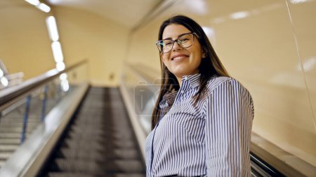 Téléchargez les photos : Jeune belle femme hispanique debout sur l'escalier roulant souriant dans la station de métro de Madrid - en image libre de droit