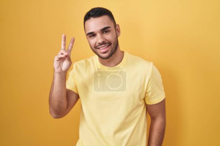 Téléchargez les photos : Jeune homme hispanique debout sur fond jaune souriant regardant la caméra montrant des doigts faisant signe de victoire. numéro deux. - en image libre de droit