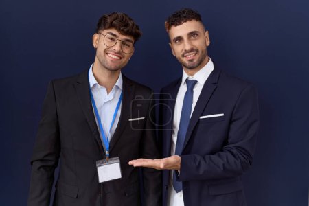 Foto de Dos hombres de negocios hispanos vistiendo ropa de negocios sonriendo alegremente presentando y señalando con la palma de la mano mirando a la cámara. - Imagen libre de derechos