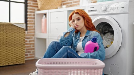 Téléchargez les photos : Jeune rousse femme tenant bouteille de détergent appuyé sur la machine à laver fatigué à la buanderie - en image libre de droit