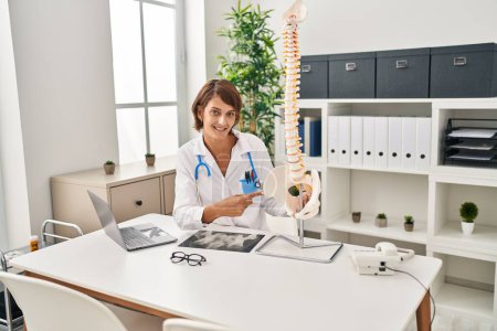 Foto de Joven hermosa doctora hispana señalando con el dedo al modelo anatómico de columna vertebral en la clínica - Imagen libre de derechos