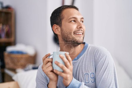 Téléchargez les photos : Young man drinking cup of coffee sitting on bed at bedroom - en image libre de droit