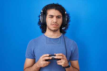 Foto de Hombre hispano con el pelo rizado jugando videojuego manteniendo controlador expresión despistado y confuso. concepto de duda. - Imagen libre de derechos