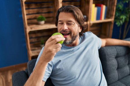 Photo for Middle age man eating apple sitting on sofa at home - Royalty Free Image