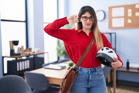 Photo for Young brunette woman working at the office holding bike helmet with angry face, negative sign showing dislike with thumbs down, rejection concept - Royalty Free Image