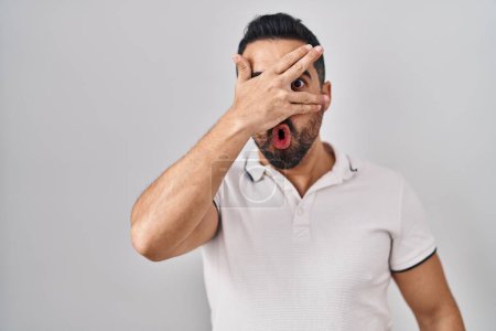 Photo for Young hispanic man with beard wearing casual clothes over white background peeking in shock covering face and eyes with hand, looking through fingers with embarrassed expression. - Royalty Free Image