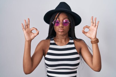 Photo for Young african american with braids wearing hat and sunglasses relaxed and smiling with eyes closed doing meditation gesture with fingers. yoga concept. - Royalty Free Image