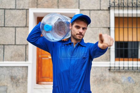 Téléchargez les photos : Jeune homme hispanique tenant une bouteille d'eau gallon pour la livraison pointant du doigt vers la caméra et vers vous, geste confiant et sérieux - en image libre de droit