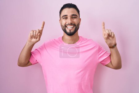 Foto de Hispanic young man standing over pink background smiling amazed and surprised and pointing up with fingers and raised arms. - Imagen libre de derechos