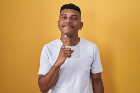 Foto de Joven hombre hispano de pie sobre fondo amarillo pensando concentrado en la duda con el dedo en la barbilla y mirando hacia arriba preguntándose - Imagen libre de derechos