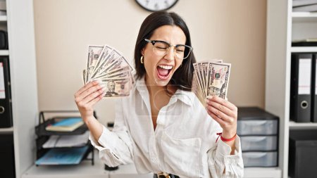 Foto de Joven hermosa mujer hispana trabajadora de negocios sonriendo confiada sosteniendo dólares en la oficina - Imagen libre de derechos