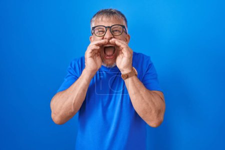 Foto de Hombre hispano con el pelo gris de pie sobre fondo azul gritando enojado en voz alta con las manos sobre la boca - Imagen libre de derechos