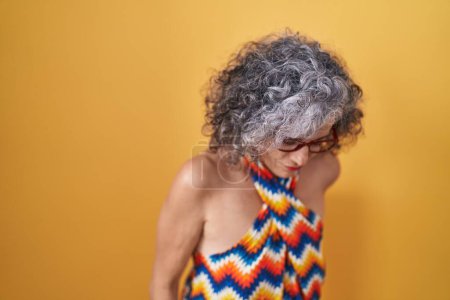 Téléchargez les photos : Femme d'âge moyen avec les cheveux gris debout sur fond jaune souffrant de maux de dos, toucher le dos avec la main, douleur musculaire - en image libre de droit