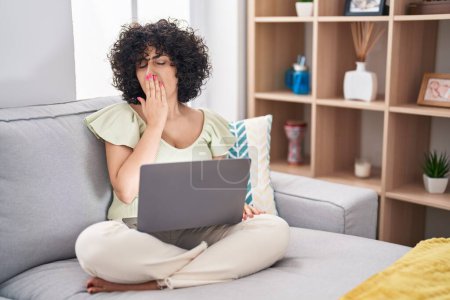 Téléchargez les photos : Jeune femme brune avec les cheveux bouclés à l'aide d'un ordinateur portable assis sur le canapé à la maison ennuyé bâillement fatigué couvrant la bouche avec la main. agitation et somnolence. - en image libre de droit