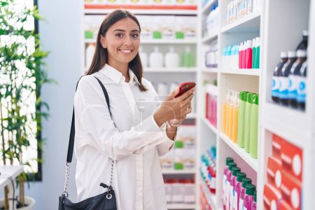 Téléchargez les photos : Young beautiful hispanic woman customer smiling confident using smartphone at pharmacy - en image libre de droit