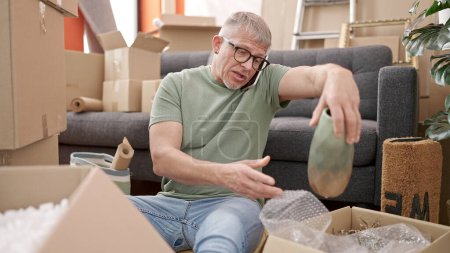 Photo for Middle age grey-haired man unpacking cardboard box talking on smartphone at new home - Royalty Free Image