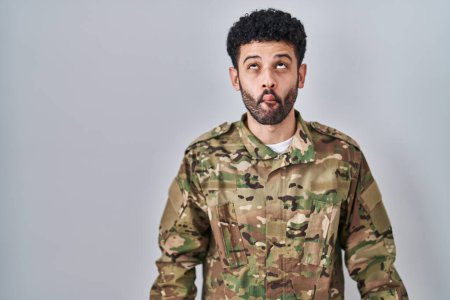 Foto de Hombre árabe vistiendo camuflaje uniforme del ejército haciendo cara de pez con labios, gesto loco y cómico. expresión divertida. - Imagen libre de derechos