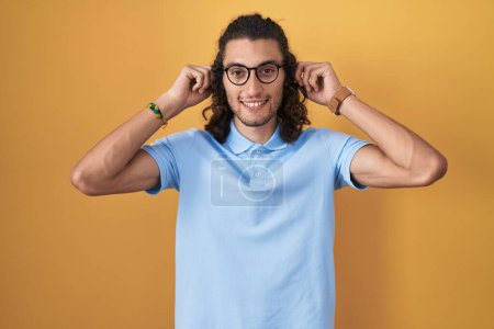 Foto de Joven hombre hispano de pie sobre fondo amarillo sonriendo tirando de las orejas con los dedos, gesto divertido. problema de audición - Imagen libre de derechos