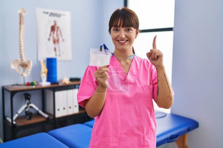 Photo for Young brunette woman working at rehabilitation clinic showing id smiling with an idea or question pointing finger with happy face, number one - Royalty Free Image