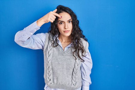 Téléchargez les photos : Jeune femme brune debout sur fond bleu pointant malheureux de boutons sur le front, infection laide de la tête noire. acné et problème de peau - en image libre de droit