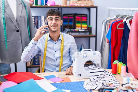 Téléchargez les photos : Jeune homme hispanique tailleur parler sur le tissu de maintien smartphone au magasin de tailleur - en image libre de droit